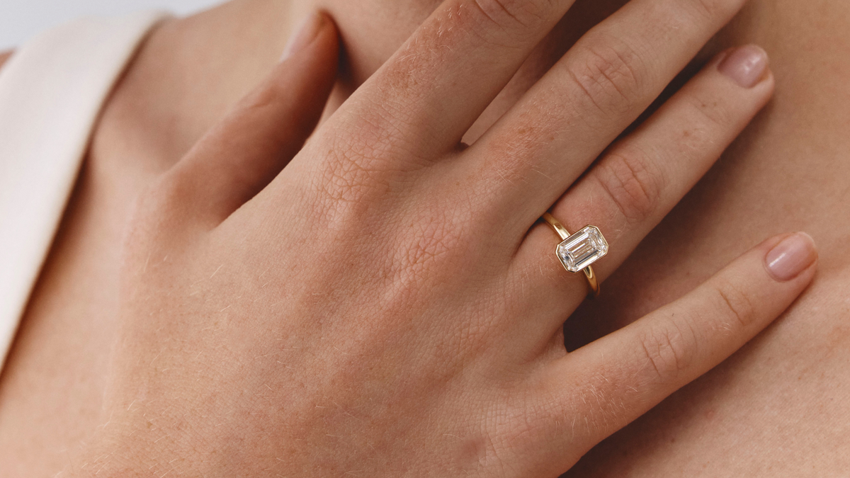 A close-up lifestyle shot of a model wearing a gold ring with an emerald-cut clear gemstone on her finger, with her hand resting on her neck against a light gray background.slider_item_dKkLm7